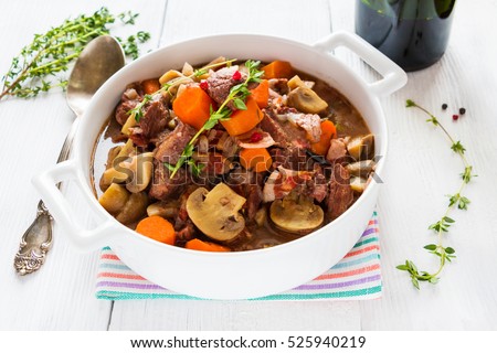 Beef Bourguignon in a casserole on white table. Stewed with bacon, garlic, carrots, onions, mushrooms,  and spices. Bunch of fresh thyme, vintage spoon, red wine.
