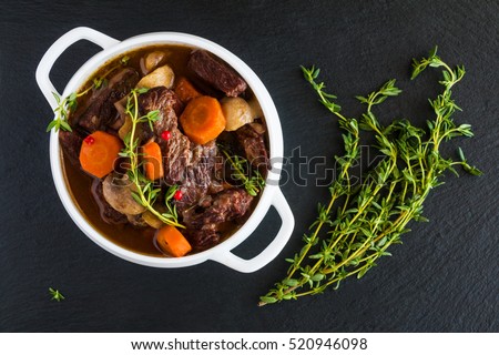 Beef Bourguignon in a white soup bowl on black stone background, top view. Stew with carrots, onions, mushrooms, bacon, garlic and bouquet garni. The dish is served with fresh thyme.