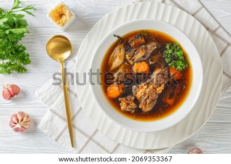 beef broth of beef meat on bones slow cooked with charred vegetables: carrot, onion, garlic, and spices served in a white bowl on a wooden table, flat lay