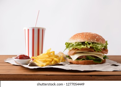 Beef Burger With Bacon And Onion Accompanied By Chips, Soda And Souces On Wooden Table With White Backgorund. Front View. Horizontal Composition.