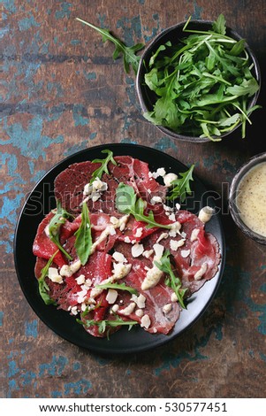 Beef carpaccio on black plate with mustard and parmesan sauce, cheese and arugula over old dark texture background. Top view with space for text