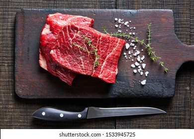 Beef cut: raw flat iron steaks of corn-fed cows on a dark wooden cutting board on a dark wooden background with fresh rosemary, sea salt, olive oil, peppercorns, garlic, view from above - Powered by Shutterstock