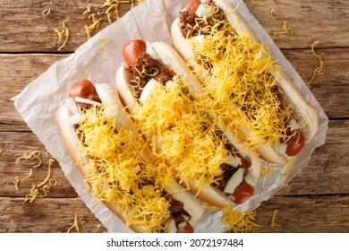 Beef hot dog in a bun covered with Cincinnati Chili, diced onions, and a mound of shredded cheddar cheese closeup in the paper on the table. Horizontal top view from above