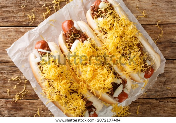 Beef hot dog in a bun\
covered with Cincinnati Chili, diced onions, and a mound of\
shredded cheddar cheese closeup in the paper on the table.\
Horizontal top view from\
above\
