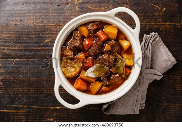 Beef meat stewed with potatoes, carrots\
and spices in ceramic pot on wooden\
background