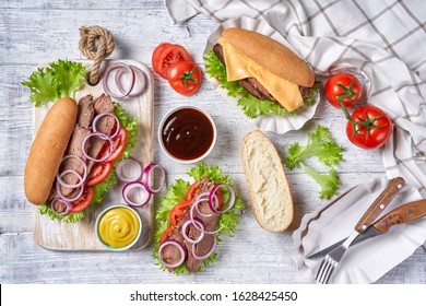 Beef Sandwich With Hoagie Rolls With Tomatoes, Cheese, Red Onion Rings, Lettuce Leaves, Served With Mustard And Bbq Sauce And Steak Cutlery On A White Wooden Table, Top View, Flat Lay