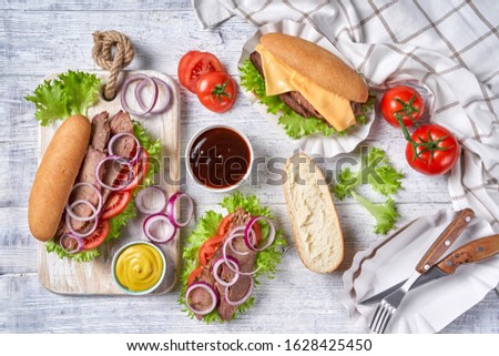 Beef sandwich with hoagie rolls with tomatoes, cheese, red onion rings, lettuce leaves, served with mustard and bbq sauce and steak cutlery on a white wooden table, top view, flat lay