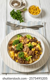 Beef Steak broccoli potato corn stew in bowl on wwhite wooden table with forks, parsley and ground black pepper, vertical view from above