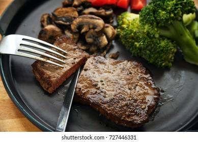 Beef Steak Is Cut With Knife And Fork, With Vegetables Like Broccoli, Mushrooms And Tomatoes, Low Carb Diet Dinner On A Dark Gray Plate, Selected Focus, Narrow Depth Of Field