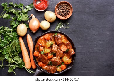 Beef stew in cast iron pan and ingredients on dark background with copy space, top view