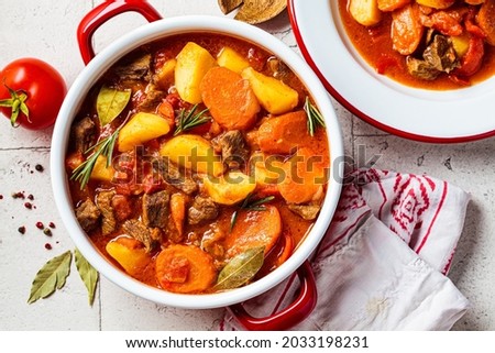 Beef stew with potatoes and carrots in tomato sauce in red pot, gray  background, top view. Slow cooking concept.