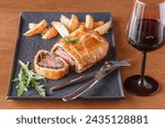 A beef wellington on a dinner plate with a side of french fries and a glass of red wine.