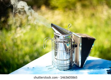 Beekeeper Smoker smokes white smoke. Apiary background. Beekeeping tool close up. Smoker stands on the ground on the green grass. Beekeeping apiary. Work at the apiary - Powered by Shutterstock