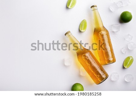 Beer advertisement and evening in pub. Two full transparent glass bottles with ale without labels, with flowing drops, lie with ice cubes and pieces of lime, on white background, free space