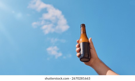 Beer bottle with blank label held against clear blue sky. Concept of packaging design, advertising, branding presentation, outdoor marketing, refreshing summer mood.
