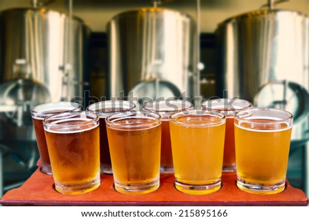 Beer flight of eight glasses of craft beer on a serving board with fermenting tanks background.
