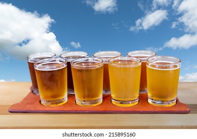 Beer Flight Of Eight Sampling Glasses Of Craft Beer On A Serving Board With Blue Cloudy Sky Background.