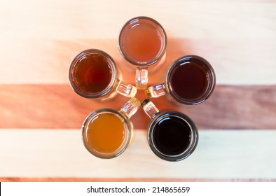 Beer Flight Of Five Sampling Mugs Of Light And Dark Craft Beer. Circle Of Little Mugs, Top View With Handles To The Center, Flower Shape.