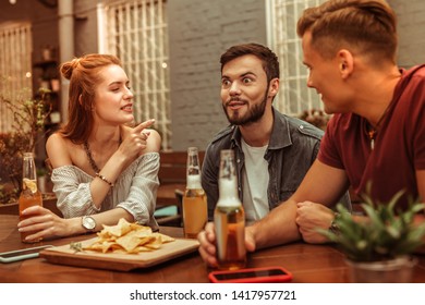 Beer with friends. Beautiful red-haired appealing young woman and two happy smiling attractive men sitting at the bar with beer