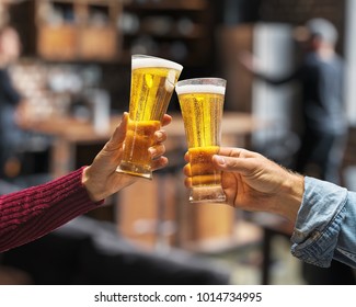 Beer glasses raised in a toast. Close-up hands with glasses. Blurred bar interior at the background.
