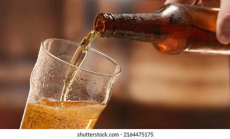 Beer is poured from dark brown bottle into beer glass. Close-up light fresh beer poured into glass steamed up from cold. Lager beer foams and pours from bottle into glass. - Shutterstock ID 2164475175