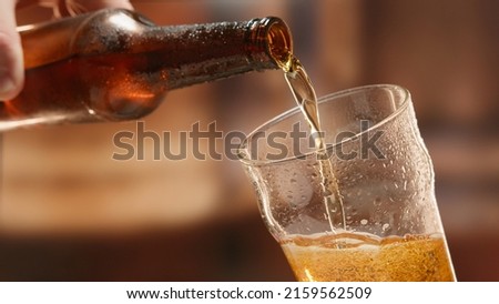 Beer is poured from dark brown bottle into beer glass. Close-up light fresh beer poured into glass steamed up from cold. Lager beer foams and pours from bottle into glass.