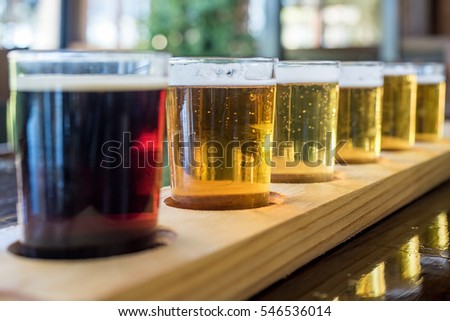Beer samplers in small glasses individually placed in holes fashioned into a unique wooden tray. Focus is on the backlit light beers, featuring some residual effervescence.