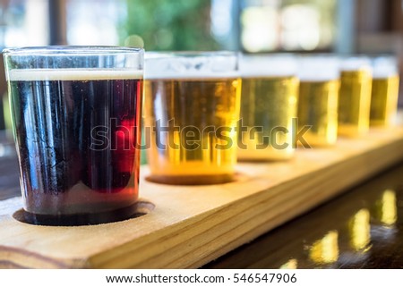 Beer samplers in small glasses individually placed in holes fashioned into a unique wooden tray. Focus is on the backlit front dark stout.