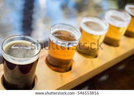 Beer samplers in small glasses individually placed in holes fashioned into a unique wooden tray. Focus is on the backlit front dark stout with shallow depth of field.