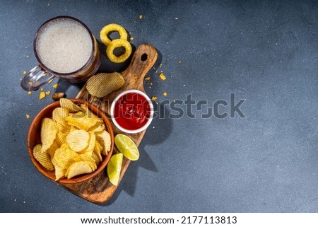 Beer with various salted snacks set. Black table background with traditional party snacks, beer bottles and glasses, with chips, onion rings, salted nuts, crisps and sauces top view copy space
