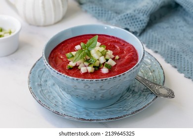 Beet and Tomato Gazpacho. Bowl of ruby red cold gaspacho puree soup topped with cucumber cubes and fresh basil on light marble background.