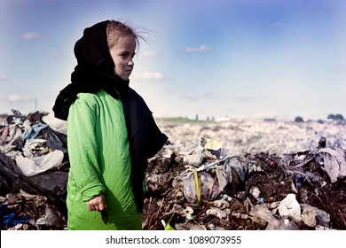 Beggar Child In Old Clothes In A Landfill Looks Sadly Forward On A Background Of Blue Sky