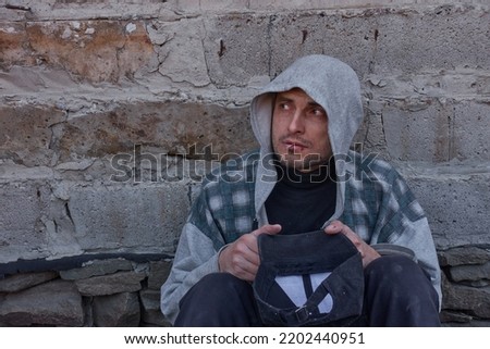 A beggar with wounded lips asks for alms. An unshaven man sits against a concrete wall, holds a baseball cap in his hand and looks sadly at people passing by.