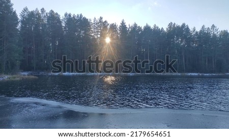 At the beginning of winter there is snow on the shores of the lake. Ice has formed in some places. The sun shines through the conifers growing on the shore. The wind drives small waves over the water
