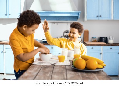 Behave Yourself. Smiling Pre-teen Boy Sitting At The Table And Raisin His Hand As If To Slap His Little Brother While He Eating Cereals With His Hands