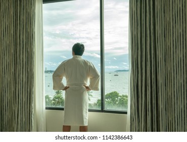 Behide Asian Man In Bathrobe Suit Open Curtain To See View Outside In Morning, Travel And Holiday Concept