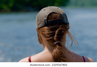 From behind, a closeup of a young woman wearing a red swimsuit and a cap on her head with her hair pulled pack in a ponytail rests while enjoying the day on a lake - Powered by Shutterstock