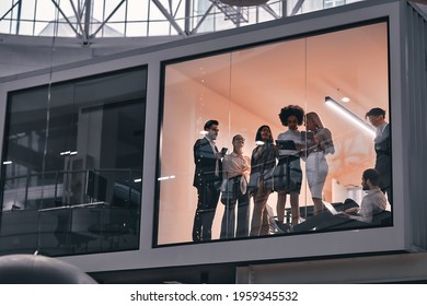 Behind The Glass In The Meeting Room In The Spacious Large Business Center, A Team Of Young People Stand And Discuss The Work.