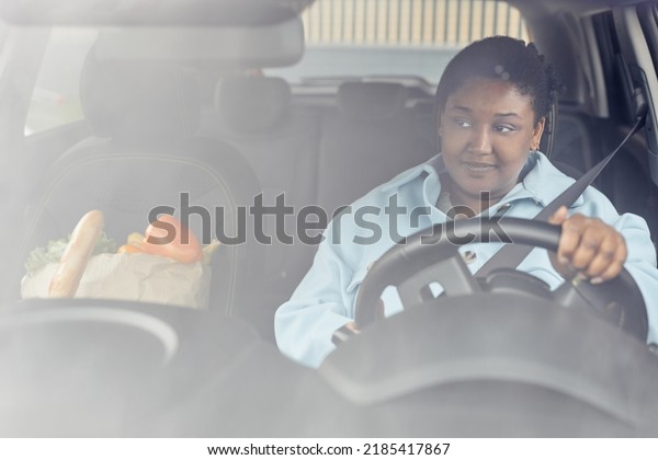 Behind glass view of\
black woman driving car with seatbelt on after shopping for\
groceries, copy space