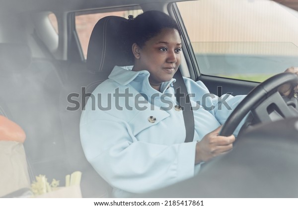 Behind glass view of young black woman driving car\
with seatbelt on, copy\
space