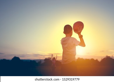 Behind Kids Holding American Football And Throwing On Sky Background On Sunset, Blurred Other Kid Children To Playing On Field