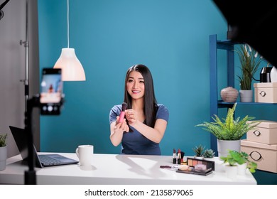 Behind the scene of asian influencer filming make up brush review using smartphone camera in studio with blue background. Vlogger creator recording cosmetics content advertising makeup product