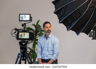 Behind The Scenes Of Man Sitting On Chair On Video Production Set In Front Of Video Camera, Tripod And Lighting Soft Box. Ready For An Interview.