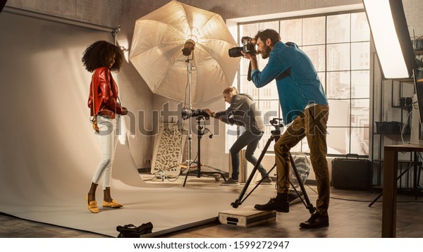 Behind the Scenes on Photo Shoot: Beautiful Black\
Model Posing for a Photographer, he Takes Photos with Professional\
Camera. Stylish Fashion Magazine Photoshoot with Pro Equipment in a\
Studio