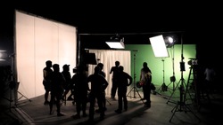 Behind The Scenes Of TV Commercial Movie Film Or Video Shooting Production Which Crew Team And Camera Man Setting Up Green Screen For Chroma Key Technique In Big Studio.