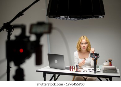 Behind The Scenes Young Blonde Woman Entrepreneur Working With Laptop Presents Cosmetic Products During Online Live Stream Over White Background Studio, Selling Online And Beauty Blogger Concept