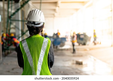 Behind a woman, a construction engineer wearing a hat and a green safety vest, stands looking at work in the construction zone.