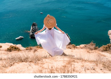 From behind, a woman is seen in a white flying dress fluttering in the wind. In a straw hat, walking down the stairs against the background of the sea with rocks and boats. The concept of travel. - Powered by Shutterstock