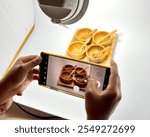 Behind-the-scenes shot of food photography, capturing hands holding a smartphone to photograph a food setup under studio lighting. Perfect for projects on photography, food styling, and creative hobbi