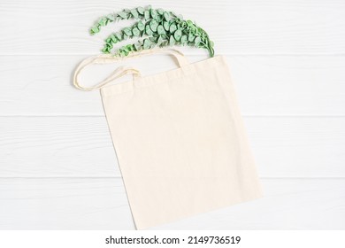 Beige Blank Template As Commercial Shopping Tote Bag Canvas And Green Eucalyptus Plant Branch On White Wood Background. Flatlay, Copyspace, Top View.